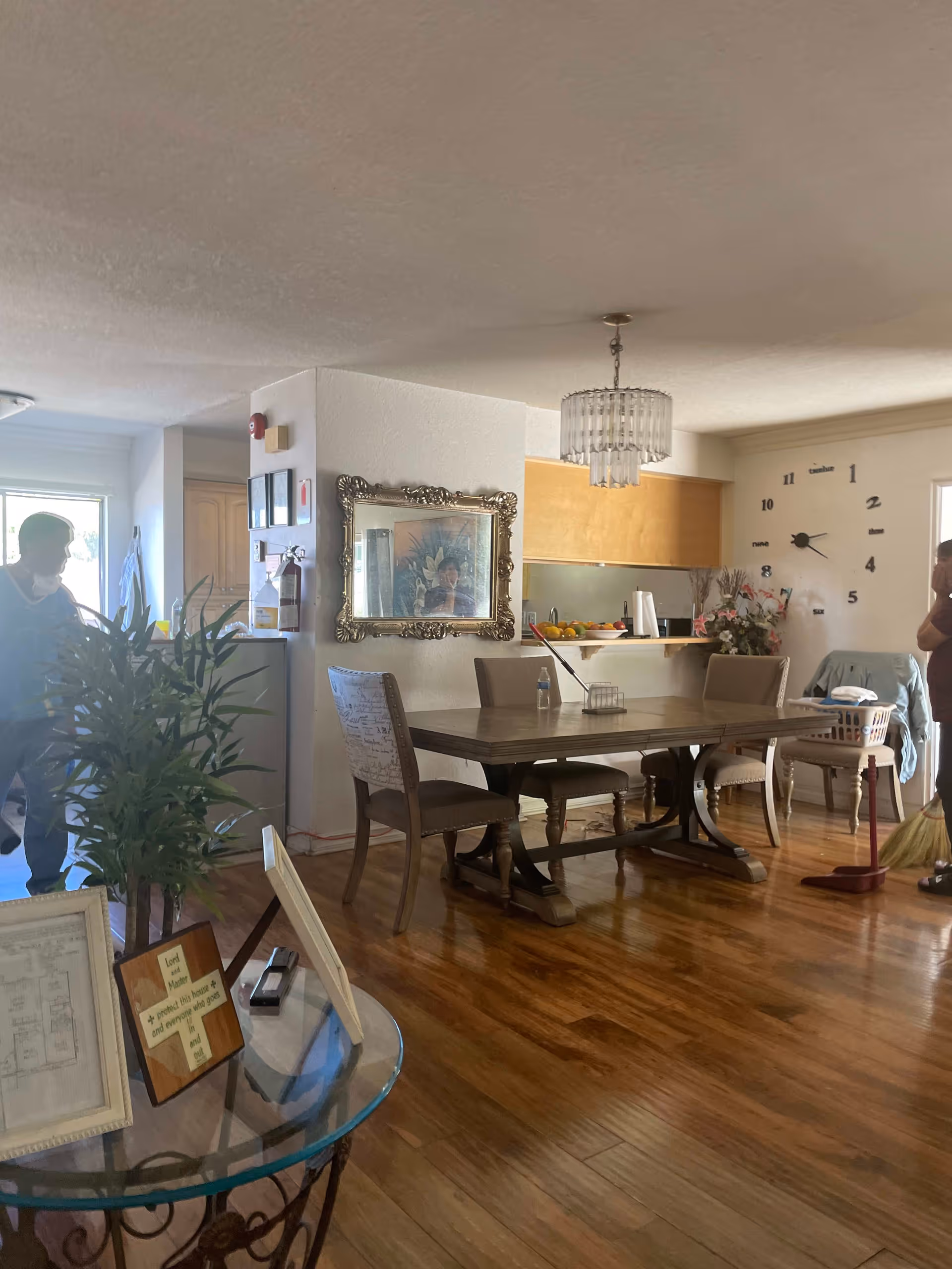 Interior view of a senior living facility dining area with a wooden dining table and chairs on a polished wooden floor. A large wall clock is visible on the far wall, along with a decorative mirror and a chandelier hanging from the ceiling. Two people are present, one near the entrance and another sweeping the floor. A glass table with framed pictures and a potted plant is in the foreground.