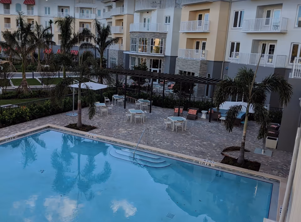 Outdoor swimming pool area at a senior living facility with palm trees, patio tables with chairs, umbrellas, and a pergola. The pool is surrounded by a paved deck and multi-story residential buildings with balconies in the background.