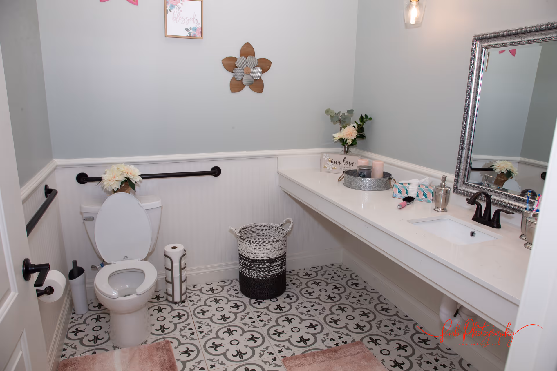 A clean and decorated bathroom with a white toilet, patterned floor tiles, a long white countertop with a sink, a large framed mirror, and decorative items including flowers, candles, and a sign. There are black grab bars on the walls and a woven basket on the floor.