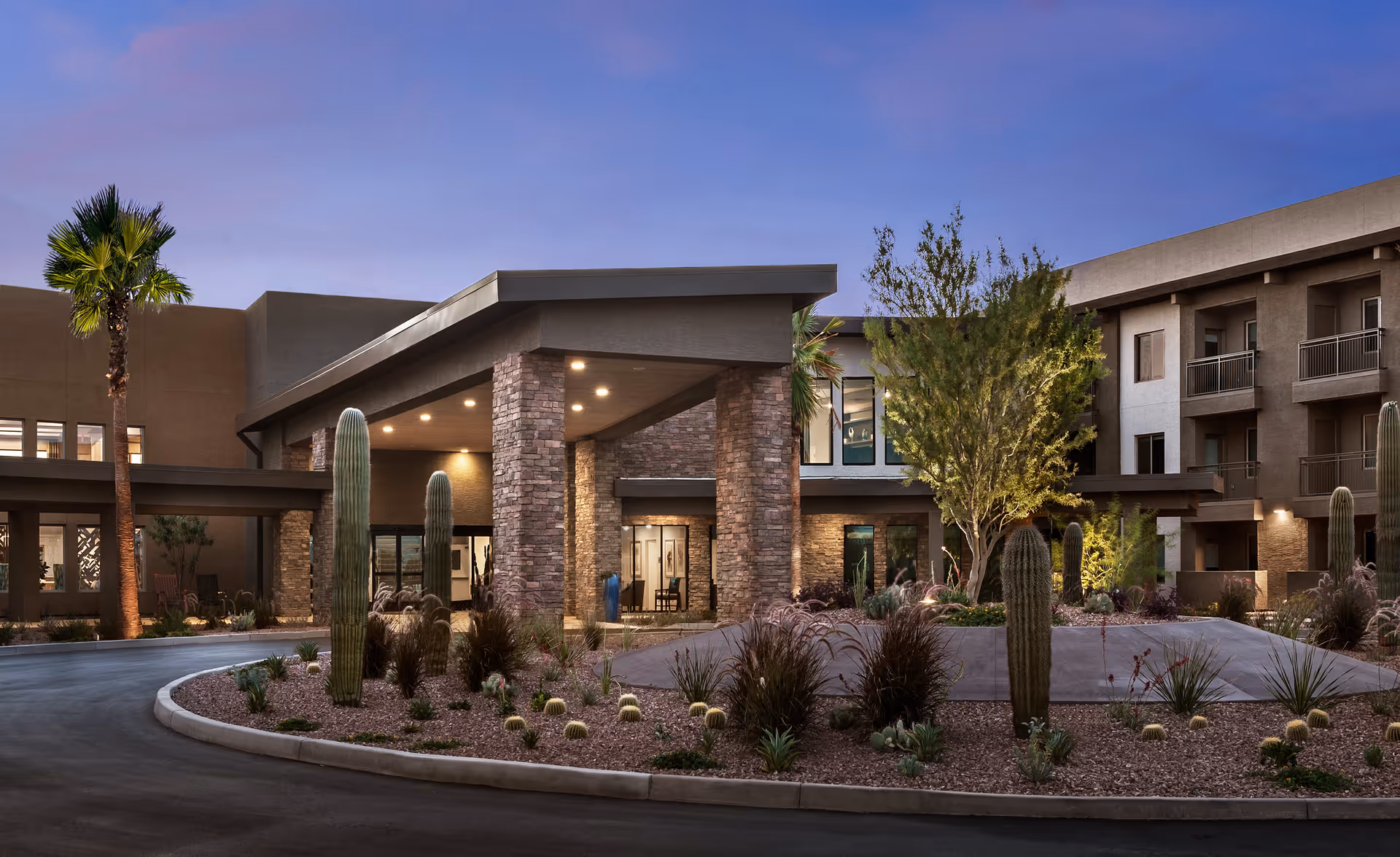Exterior view of Inspira at Arrowhead senior living facility at dusk, featuring a modern building entrance with stone pillars, desert landscaping including cacti and palm trees, and a curved driveway.