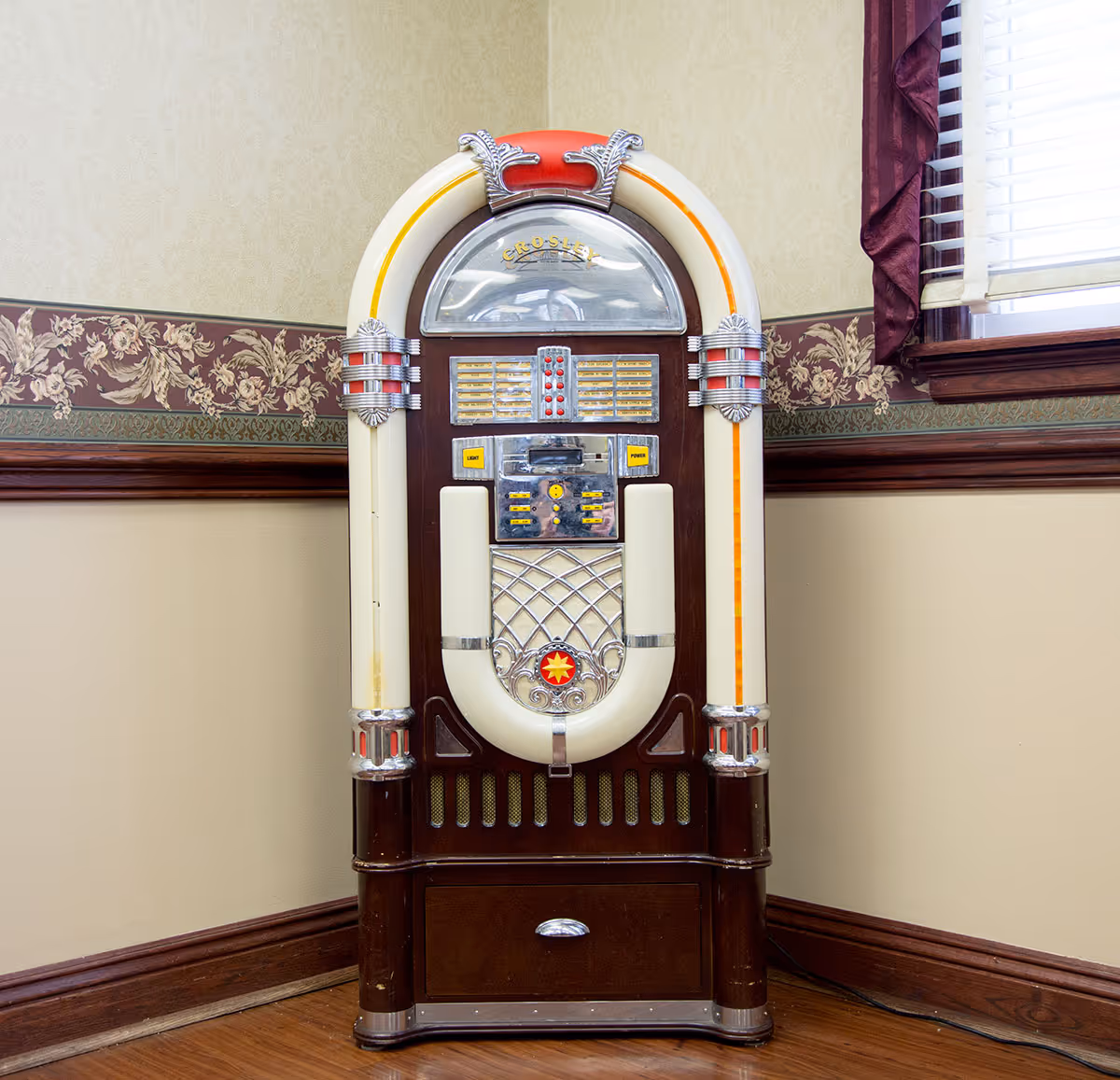 A vintage-style jukebox with a wooden base, cream and red accents, and chrome details, placed in the corner of a room with floral wallpaper border and a window with blinds and burgundy curtains.