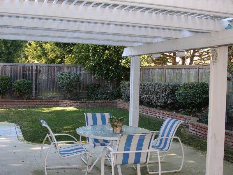 Outdoor patio area with a white pergola overhead, a round glass table with four white metal chairs featuring blue and white striped cushions. The patio overlooks a well-maintained green lawn bordered by a wooden fence and trimmed bushes and shrubs.