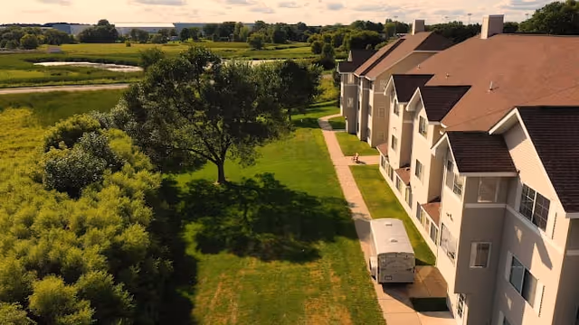 Aerial view of a multi-story assisted living facility building with a brown roof and beige exterior walls, adjacent to a green lawn with trees and shrubs. A paved walkway runs alongside the building, and a vehicle is parked near the entrance. The background features open fields and a partly cloudy sky.