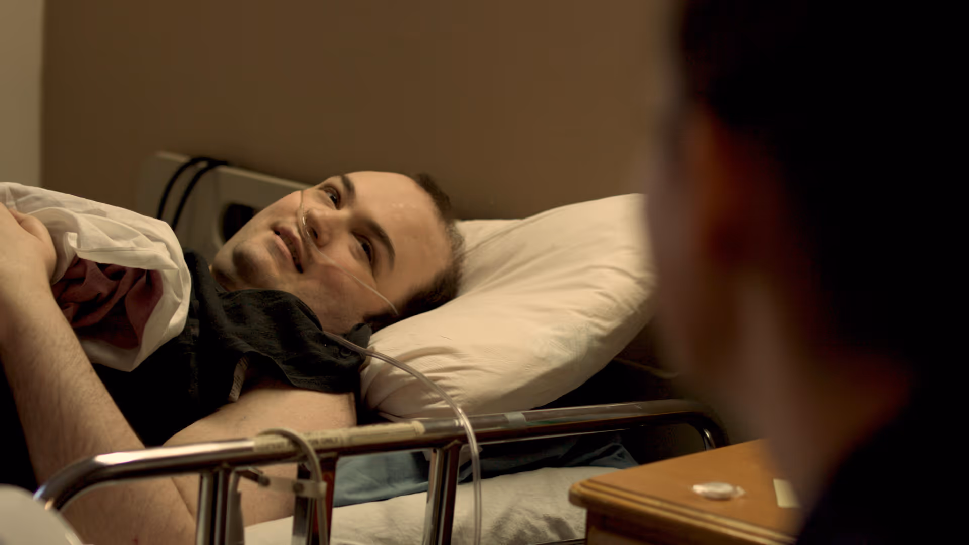 A man lying in a hospital bed with an oxygen tube in his nose, looking and smiling at another person who is partially visible in the foreground.