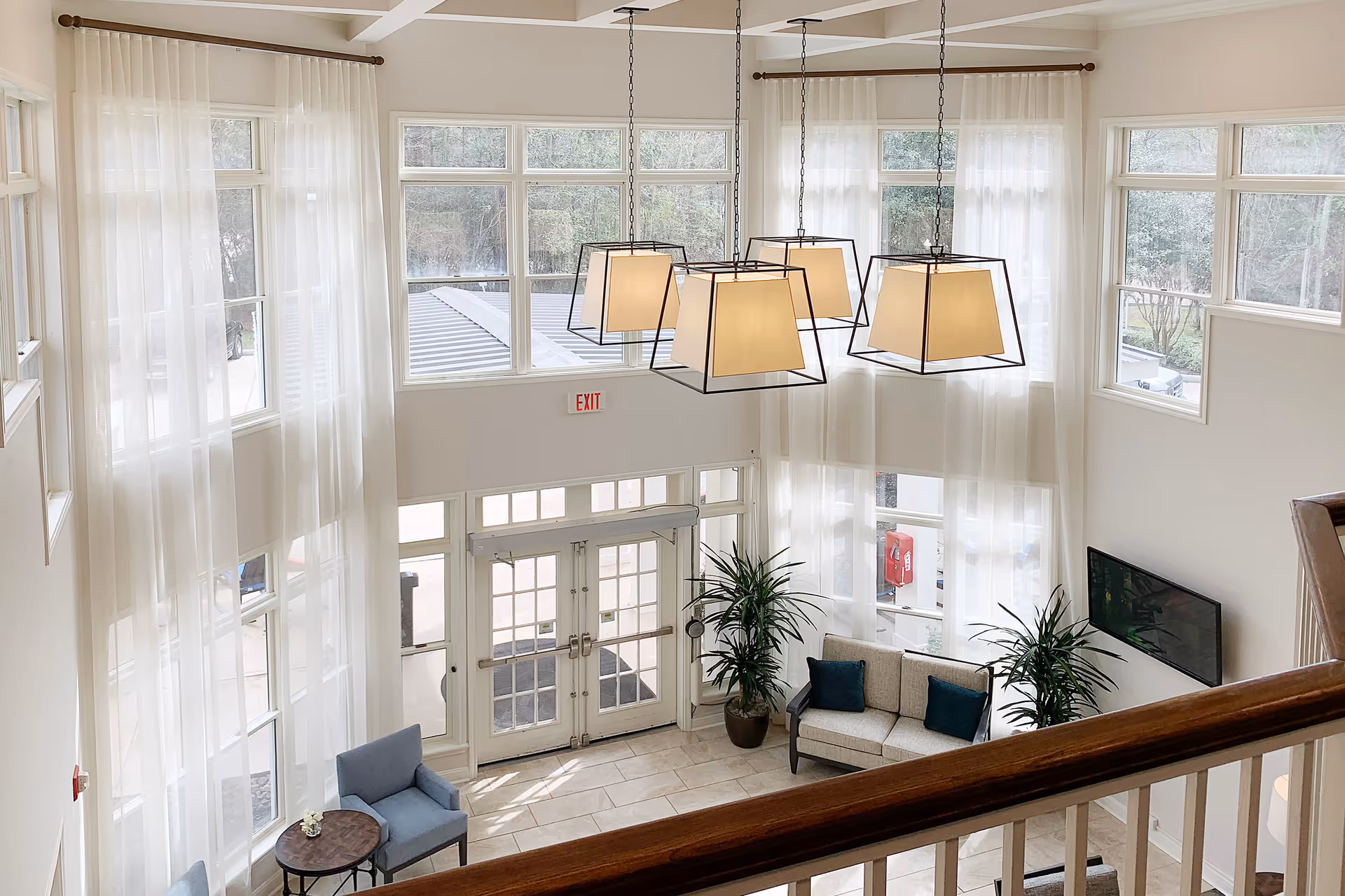 Bright double-height lobby with large windows, hanging pendant lights, seating areas and glass entrance doors.