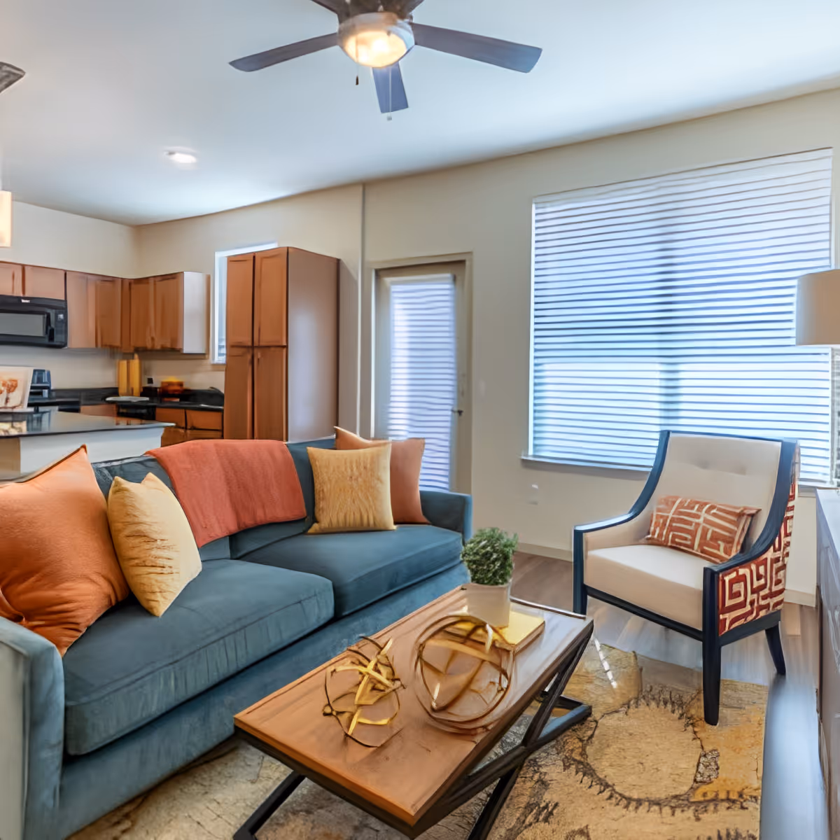 A modern living room with a blue sofa adorned with orange and yellow pillows and a matching orange throw blanket. There is a wooden coffee table with decorative golden orbs and a small potted plant. To the right, there is a white armchair with dark wooden arms and legs, featuring a patterned orange cushion. The room has large windows with horizontal blinds, a ceiling fan with a light, and an open view into a kitchen area with wooden cabinets and black appliances.