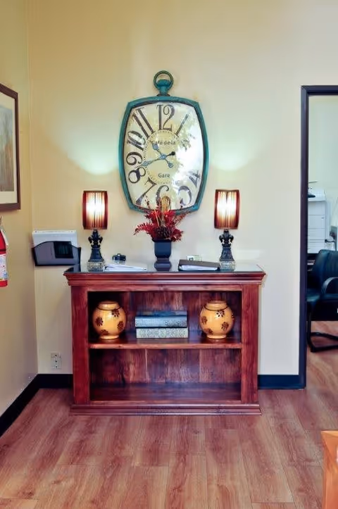 A wooden console table with two decorative vases and three books on the lower shelf, two table lamps with striped shades on top, and a floral arrangement in the center. Above the table is a large vintage-style wall clock with distorted numbers. The room has light-colored walls and wood flooring, with a doorway leading to another room with office chairs visible.