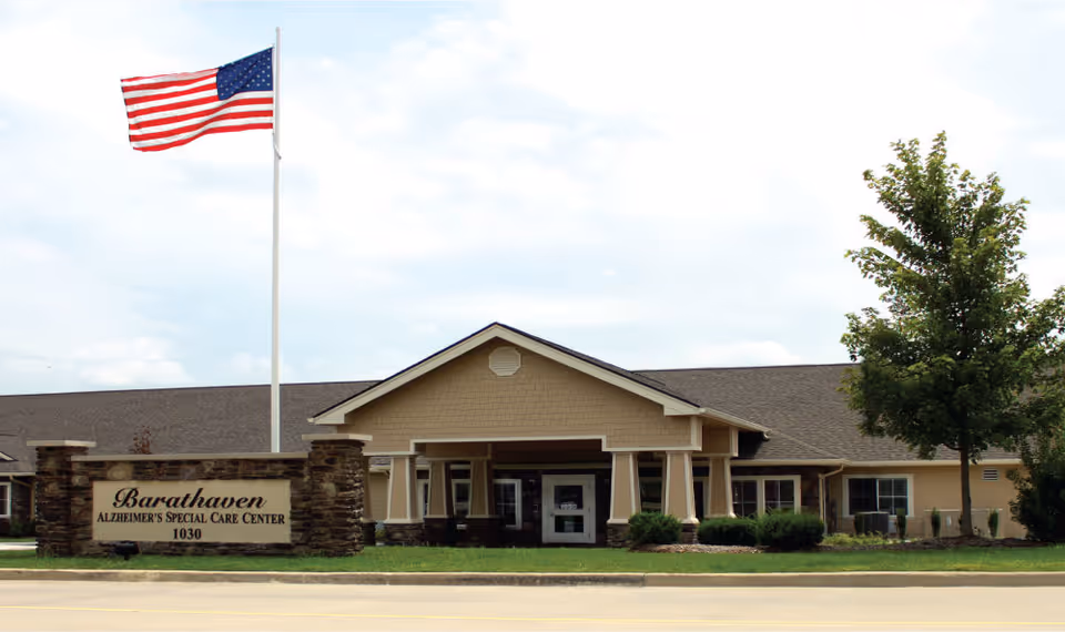 Exterior front view of Barathaven Alzheimer's Special Care Center building with a large American flag on a flagpole and a sign displaying the facility name and address in front of the building.
