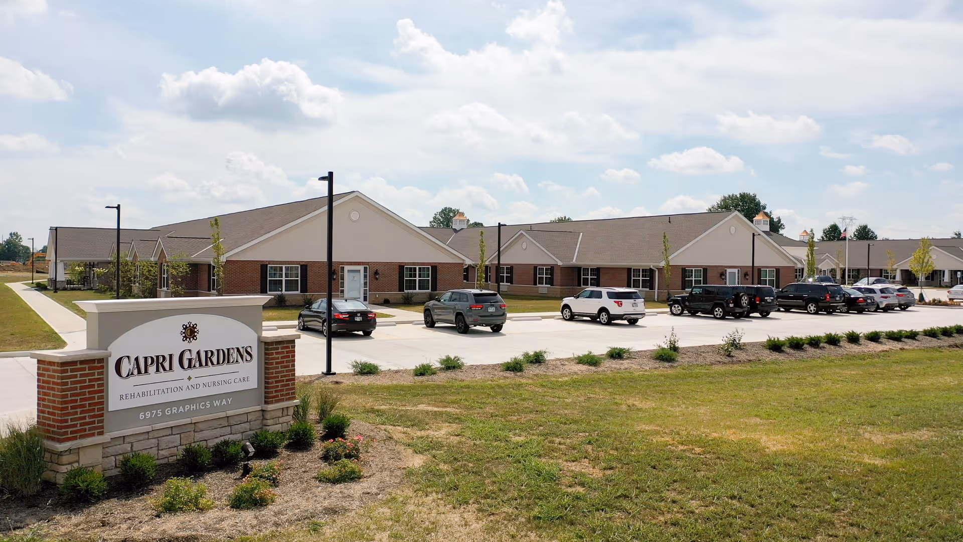 Exterior view of Capri Gardens Nursing Center showing a single-story brick building with multiple windows and a parking lot with several cars. The sky is partly cloudy and there is a landscaped area with grass and small plants in the foreground. A sign in front reads 'Capri Gardens Rehabilitation and Nursing Care, 6975 Graphics Way.'