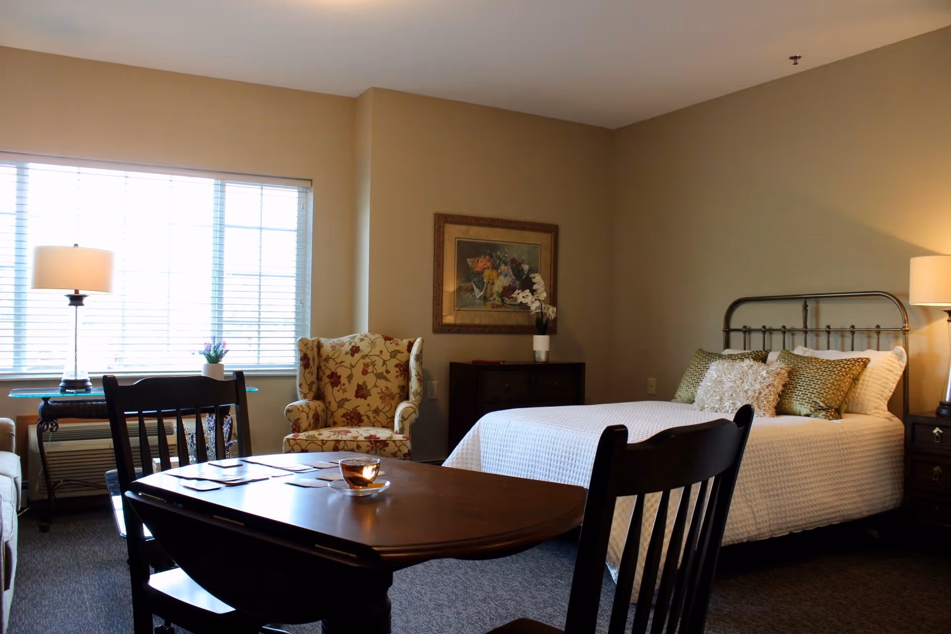 Well-lit furnished bedroom with a bed, floral armchair, table and chairs, and lamps by a large window.