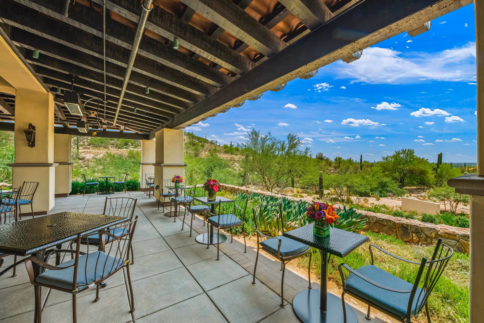 Outdoor covered patio area with several small tables and metal chairs with blue cushions, overlooking a scenic desert landscape with green shrubs, cacti, and a bright blue sky with scattered clouds.