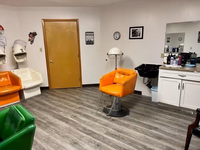 Interior of a salon area with orange, green, and white salon chairs, hair dryers mounted on the wall, a wooden door, a clock, a framed picture, a black sink, and white cabinets with various hair products on the counter.