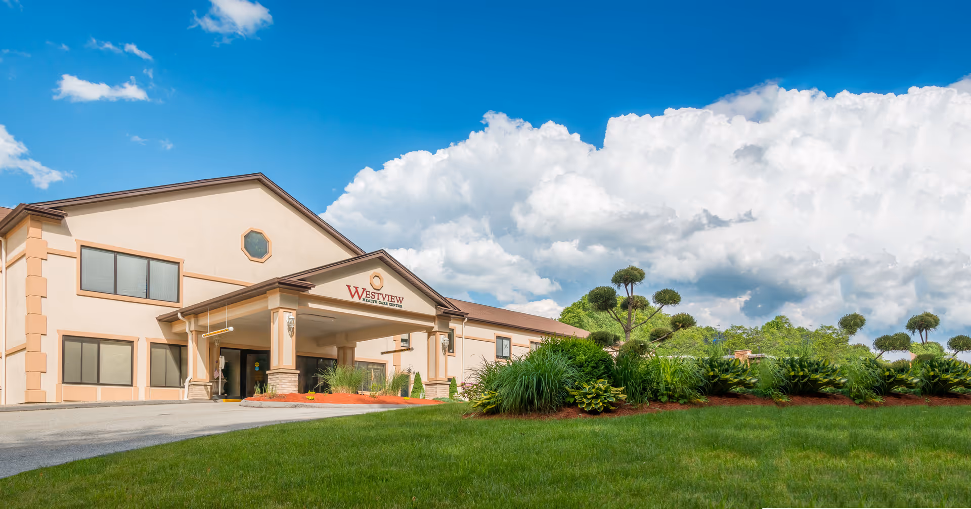 Exterior view of Westview Health Care Center building with a beige facade, brown roof, and a covered entrance. The building is surrounded by well-maintained green grass, landscaped bushes, and uniquely trimmed trees under a bright blue sky with large white clouds.