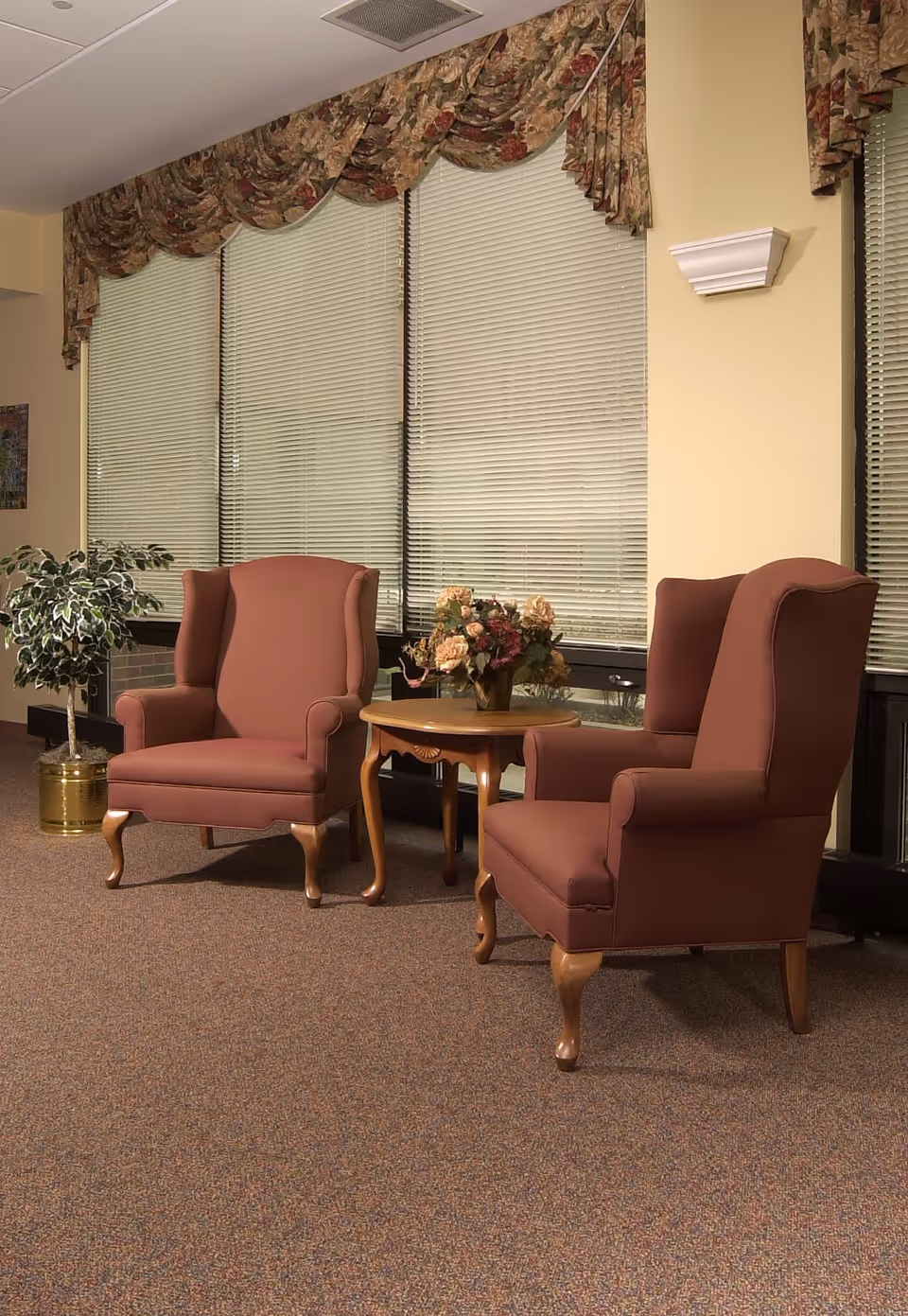 Two upholstered armchairs with wooden legs positioned around a small wooden table with a floral arrangement, set in a room with large windows covered by blinds and floral valances, a potted plant in a brass container, and beige walls.