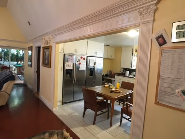 View of a residential kitchen and dining area with a wooden table and four chairs. The kitchen features white cabinets, a stainless steel refrigerator, and various kitchen appliances. The dining area is adjacent to a hallway with wooden flooring and a glimpse of a living room with a couch. A dog is partially visible on the floor in the foreground.