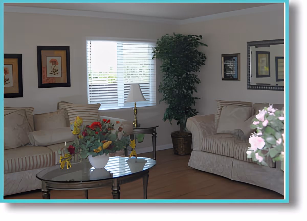 A cozy living room with two beige sofas adorned with cushions, a glass coffee table with a flower arrangement, a side table with a lamp, a large potted plant in the corner, framed artwork on the walls, and a window with blinds letting in natural light.