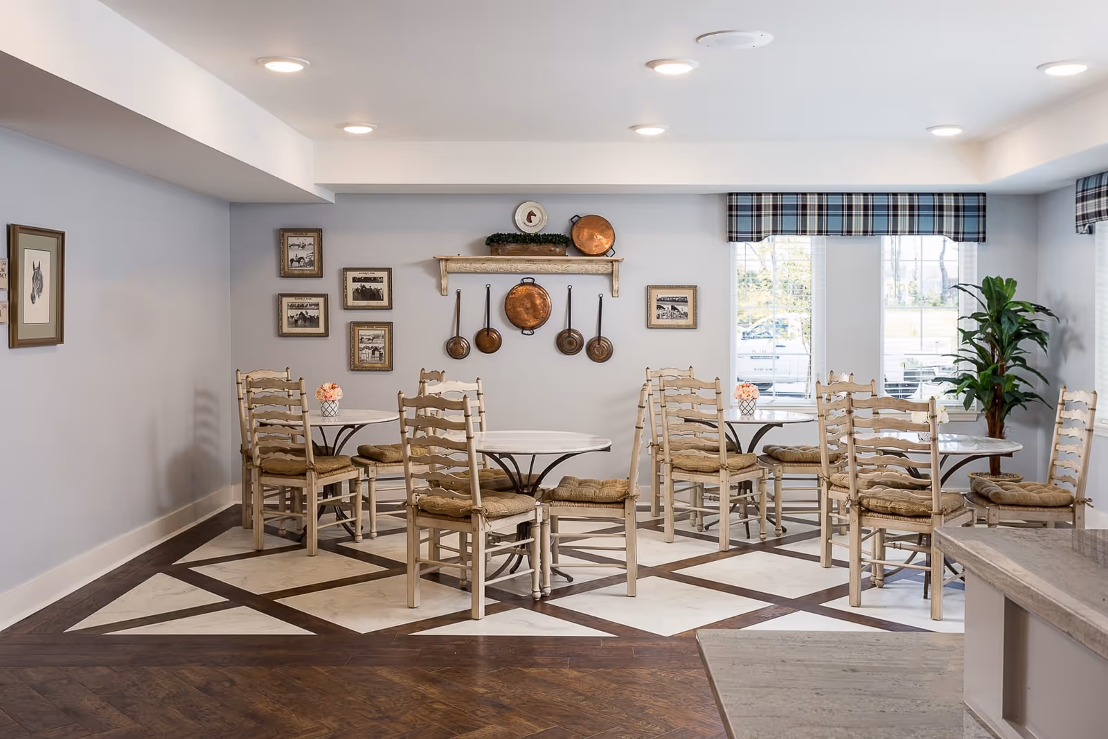 A cozy dining area with several round tables and wooden chairs with cushions. The walls are decorated with framed pictures and hanging copper pans. Large windows with plaid valances let in natural light, and there is a green potted plant in the corner.