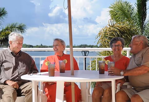 Four elderly people sitting around a white outdoor table with drinks, enjoying a sunny day near a body of water with palm trees in the background.