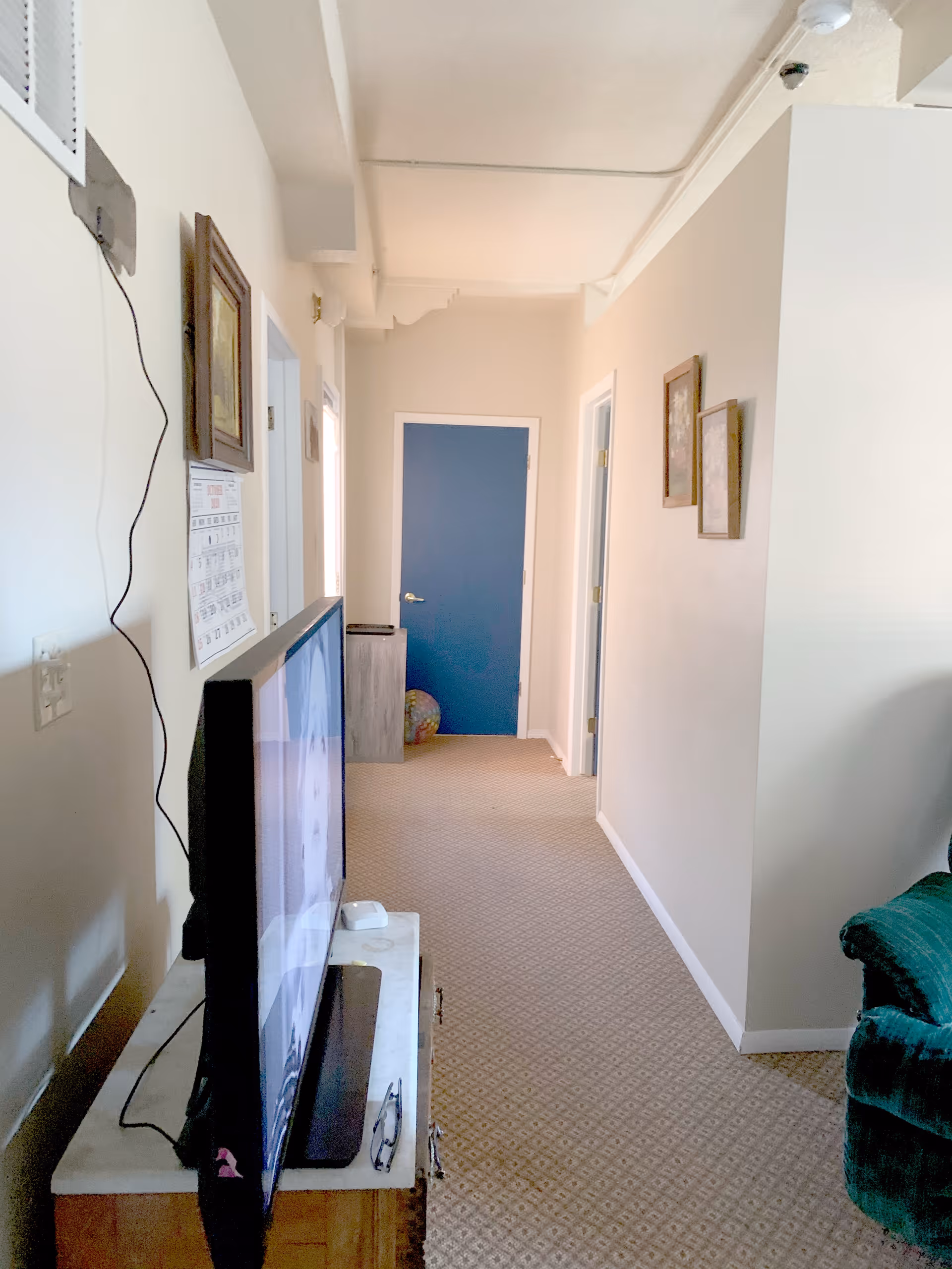 Interior hallway of a senior living facility with beige walls and carpeted floor. A flat-screen TV is placed on a wooden stand on the left side, and a green upholstered chair is partially visible on the right. The hallway leads to a blue door at the end, with two additional doors on the right side. There are framed pictures hanging on the right wall.