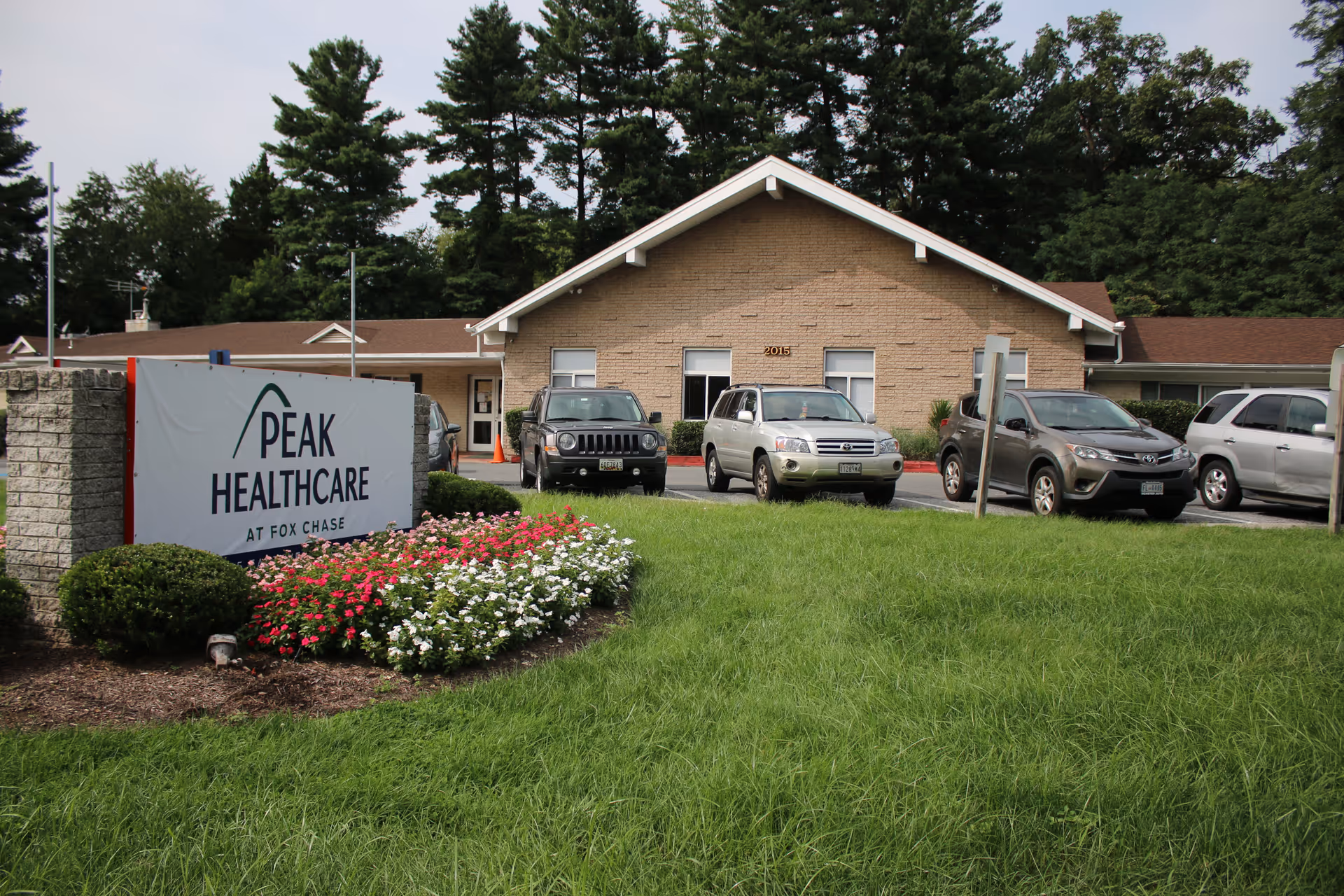Exterior view of a healthcare facility building with a sign that reads 'Peak Healthcare at Fox Chase' in front. There are several parked cars and a well-maintained lawn with flower beds. Trees are visible in the background.