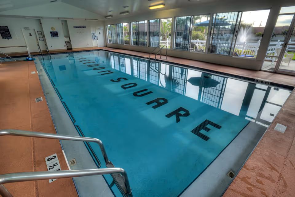 Indoor swimming pool with the words 'FREEDOM SQUARE' visible on the pool floor. The pool area has large windows showing an outdoor patio with umbrellas and a fountain. There are metal handrails and depth markers around the pool.