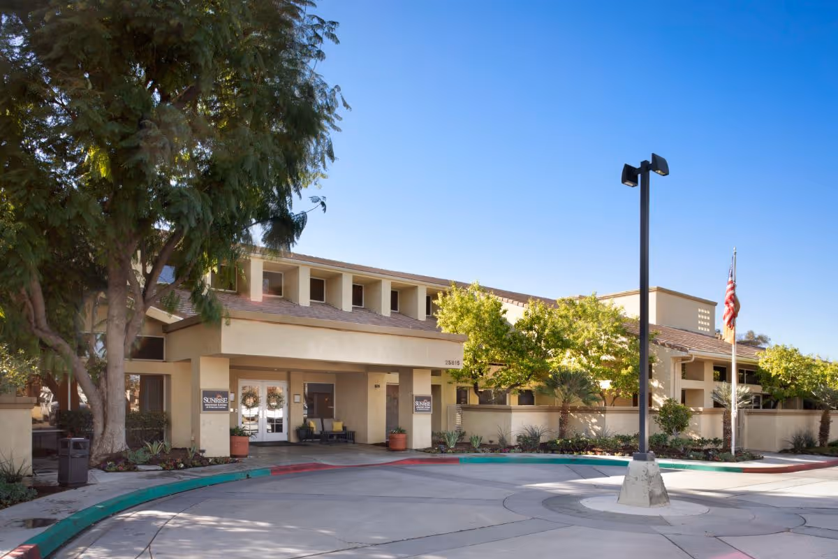 Exterior view of Sunrise At Sterling Canyon facility showing the entrance with a covered driveway, trees, and landscaping under a clear blue sky.