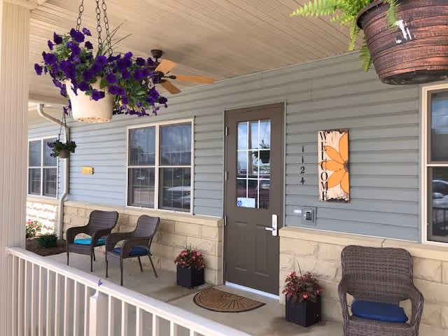 Covered porch area at the entrance of a building with light blue siding and stone accents. There are three wicker chairs with blue cushions, two black planters with red flowers, and hanging flower pots with purple and green plants. A ceiling fan is mounted on the porch ceiling. The door is brown with a glass window and a welcome sign with a large orange flower is mounted on the wall next to it. The number 124 is displayed beside the door.