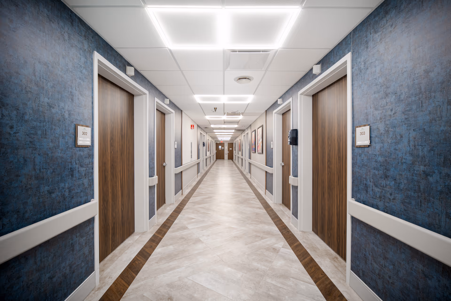A long, well-lit hallway in a healthcare facility with blue textured walls, wooden doors on both sides, white handrails, and framed artwork along the right wall.