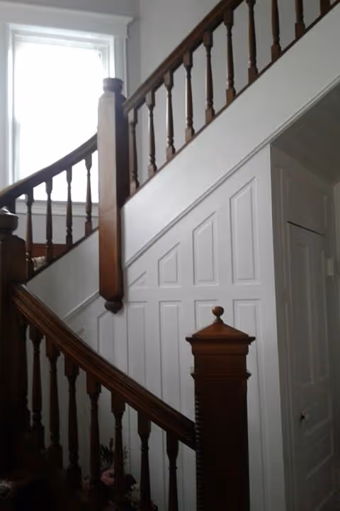 Interior view of a staircase with wooden handrails and white paneled walls. A window with white trim is visible at the top of the stairs, allowing natural light to enter.