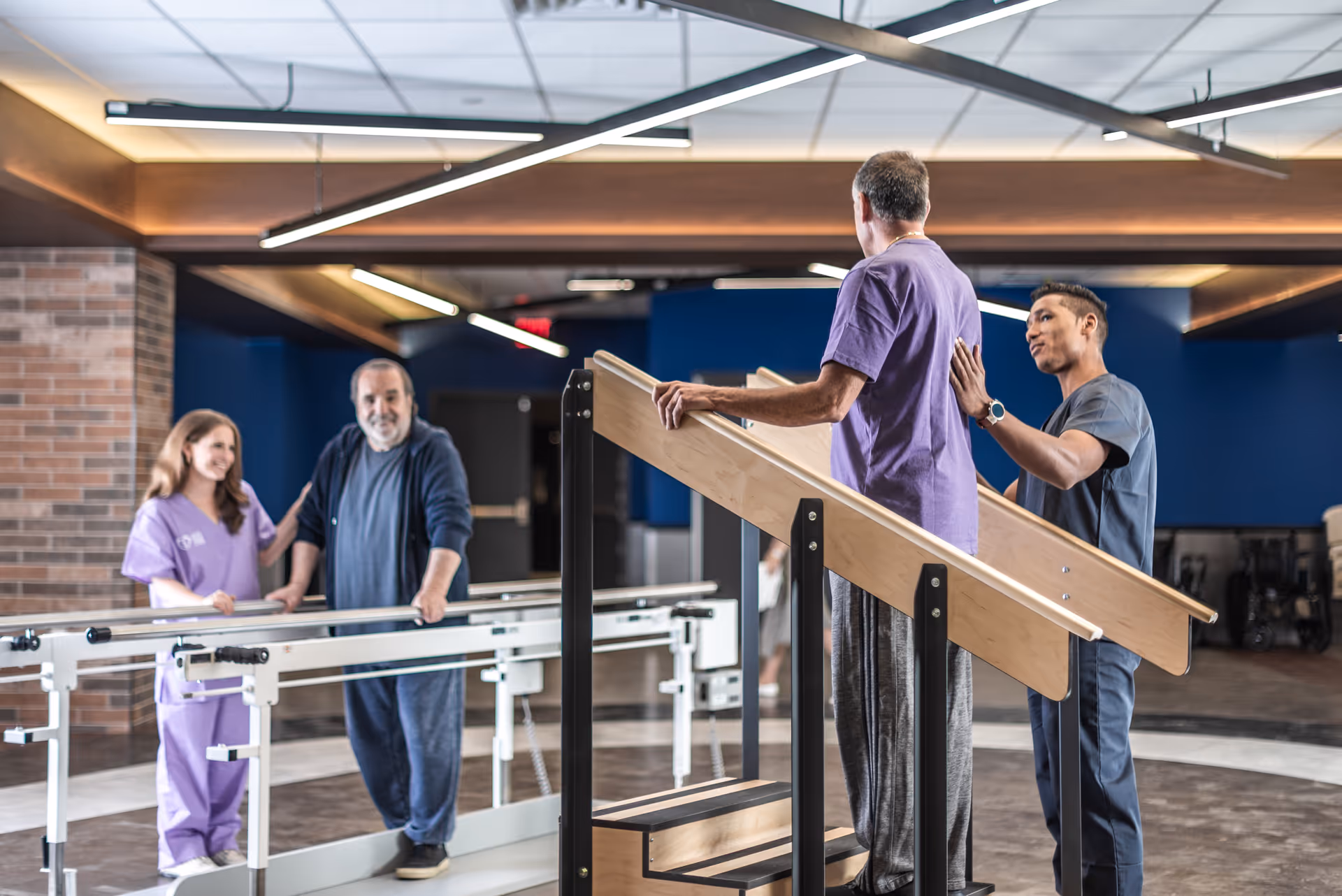 A rehabilitation room where two elderly men are assisted by two healthcare professionals. One elderly man is walking on parallel bars with the support of a female healthcare worker in purple scrubs. The other elderly man is walking up wooden stairs with handrails, assisted by a male healthcare worker in dark scrubs. The room has modern lighting and a blue and brick wall background.
