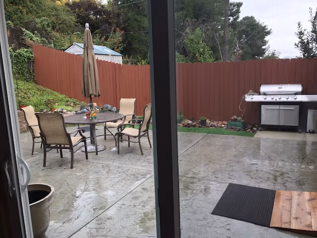 Outdoor patio area with a round table and six chairs, a closed umbrella in the center of the table, a barbecue grill against a red wooden fence, and some greenery and trees in the background. The patio surface appears wet, possibly from recent rain.