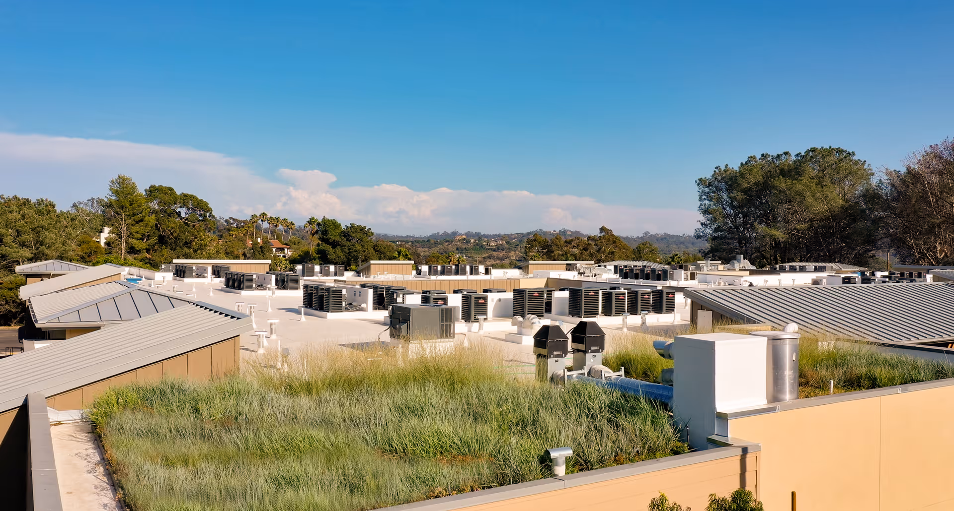 View of the rooftop of a building at Westmont of Encinitas with HVAC units and a section of green roof with grass. Trees and hills are visible in the background under a clear blue sky.
