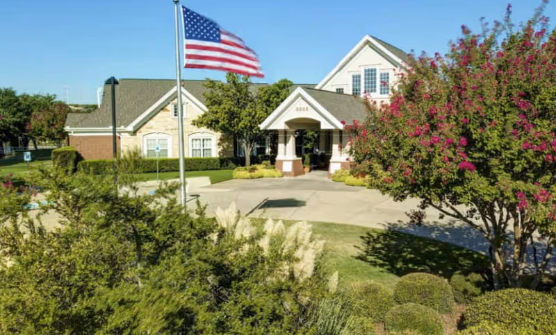 Front entrance of a two-story residential-style building with a flagpole, circular driveway, and landscaped shrubs and trees.