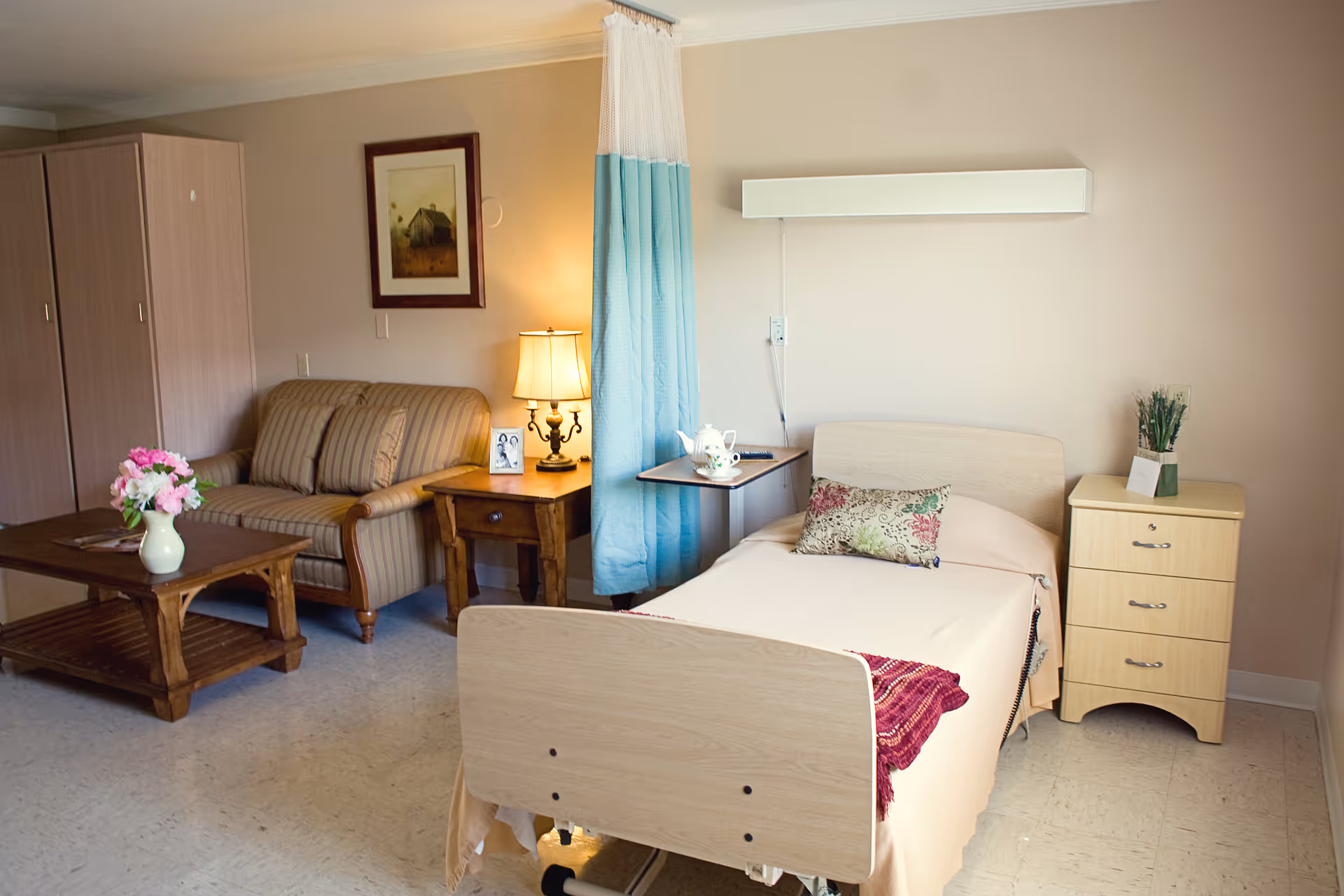 A well-lit senior living facility room featuring a single hospital-style bed with a floral pillow and a red throw blanket. Next to the bed is a small wooden nightstand with a potted plant and a wall-mounted light fixture above. A blue privacy curtain partially separates the bed area from a sitting area that includes a striped loveseat, a wooden side table with a lamp and a framed photo, and a wooden coffee table with a vase of pink flowers. A framed picture hangs on the wall above the loveseat.