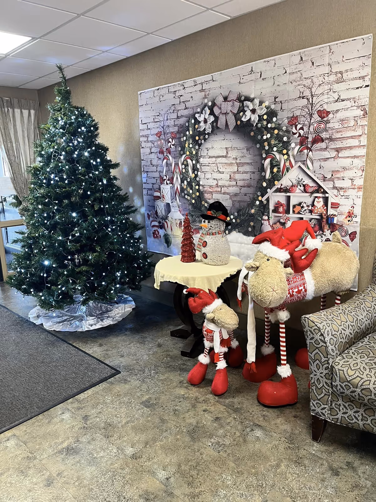 A decorated indoor corner with a Christmas tree adorned with white lights, a small round table covered with a yellow cloth holding a red Christmas tree decoration and a snowman figure. Next to the table are two plush reindeer figures wearing red and white Christmas outfits. Behind the table is a large festive wall decoration featuring a wreath and candy-themed ornaments. A patterned armchair is partially visible on the right side.