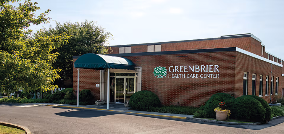 Exterior view of a brick building with a green awning over the entrance. The building has a sign that reads 'GREENBRIER HEALTH CARE CENTER'. There are bushes and a tree near the entrance, and a paved driveway in front.