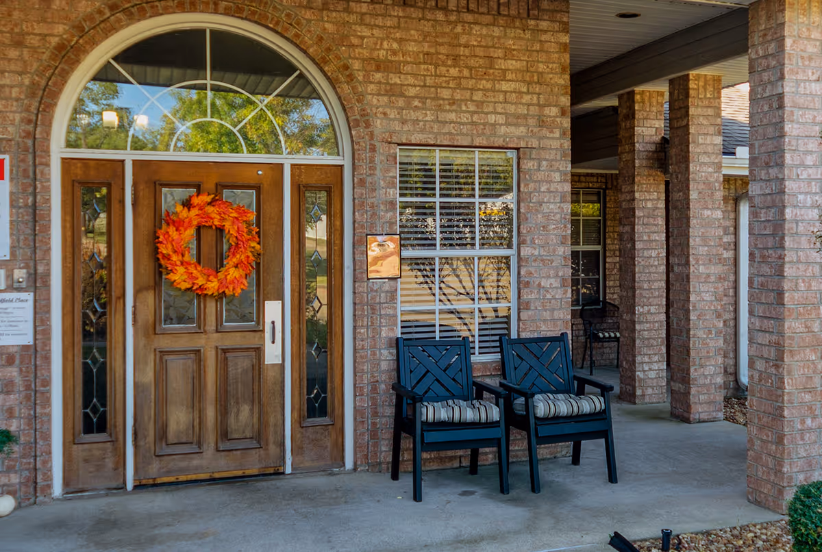 Front entrance of a brick building with a wooden door decorated with an orange and red autumn leaf wreath. Two black chairs with striped cushions are placed on the porch next to a window with white blinds. The porch is supported by brick columns.