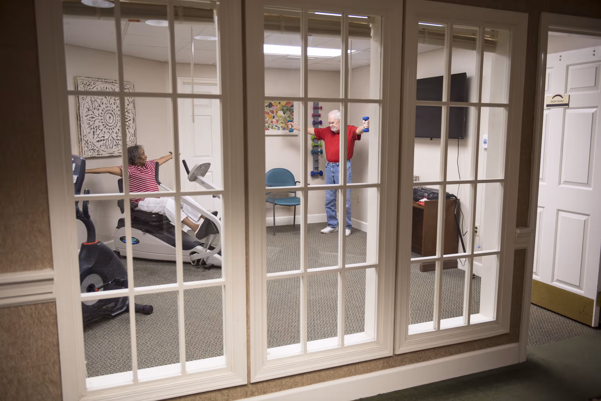 View through paned glass into a small exercise room where two residents use fitness equipment and hand weights.