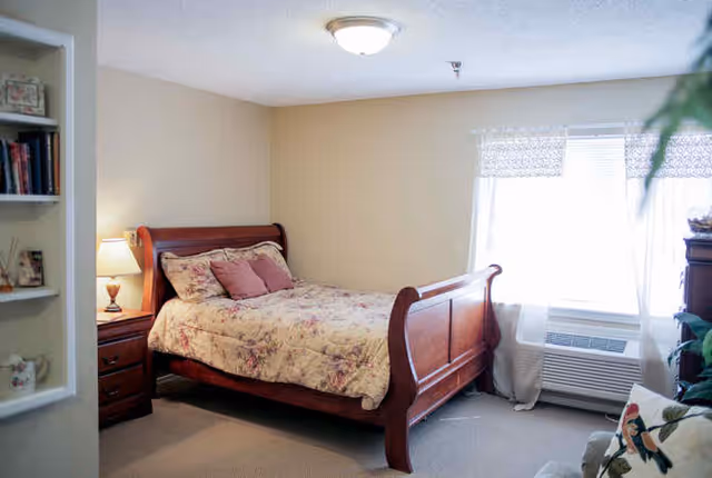 A cozy bedroom with a wooden sleigh bed covered in floral bedding and several pillows. There is a wooden nightstand with a lamp beside the bed. A window with sheer curtains lets in natural light, and an air conditioning unit is installed below the window. A bookshelf with books and decorative items is partially visible on the left, and a cushioned chair with a floral pillow is on the right.