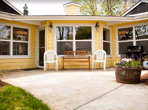 Outdoor patio area of a yellow building with a wooden bench flanked by two white plastic chairs. There is a large window behind the bench, two wall-mounted lantern lights, a potted plant in a wooden barrel on the right, and a barbecue grill nearby. Green grass and trees are visible in the background.