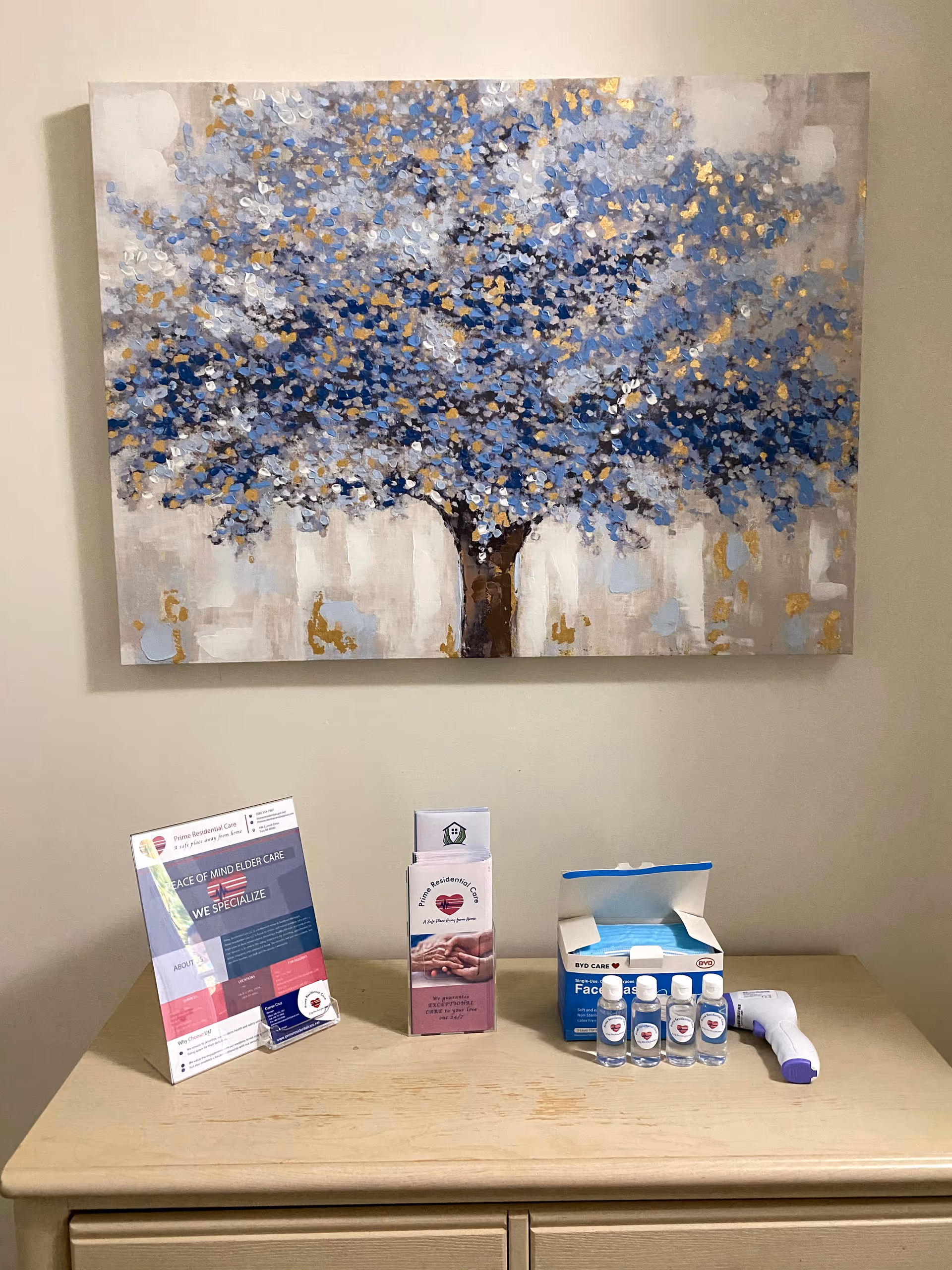 A beige wooden dresser with informational brochures, small bottles of hand sanitizer, a box of face masks, and a digital thermometer on top. Above the dresser hangs a painting of a tree with blue and gold leaves on a neutral background.
