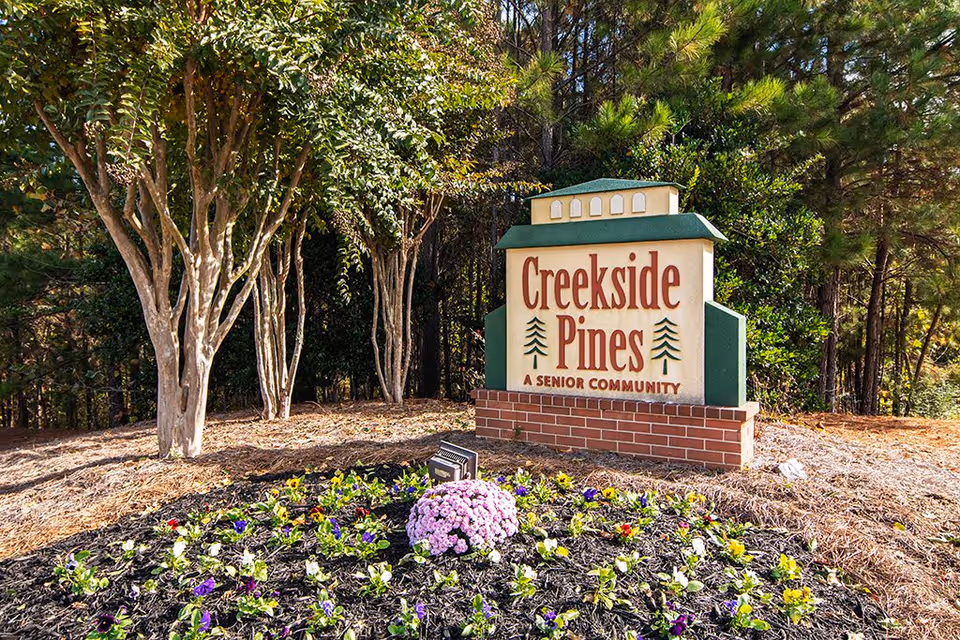 Outdoor sign for Creekside Pines, a senior community, surrounded by trees and a flower bed with colorful flowers in front.