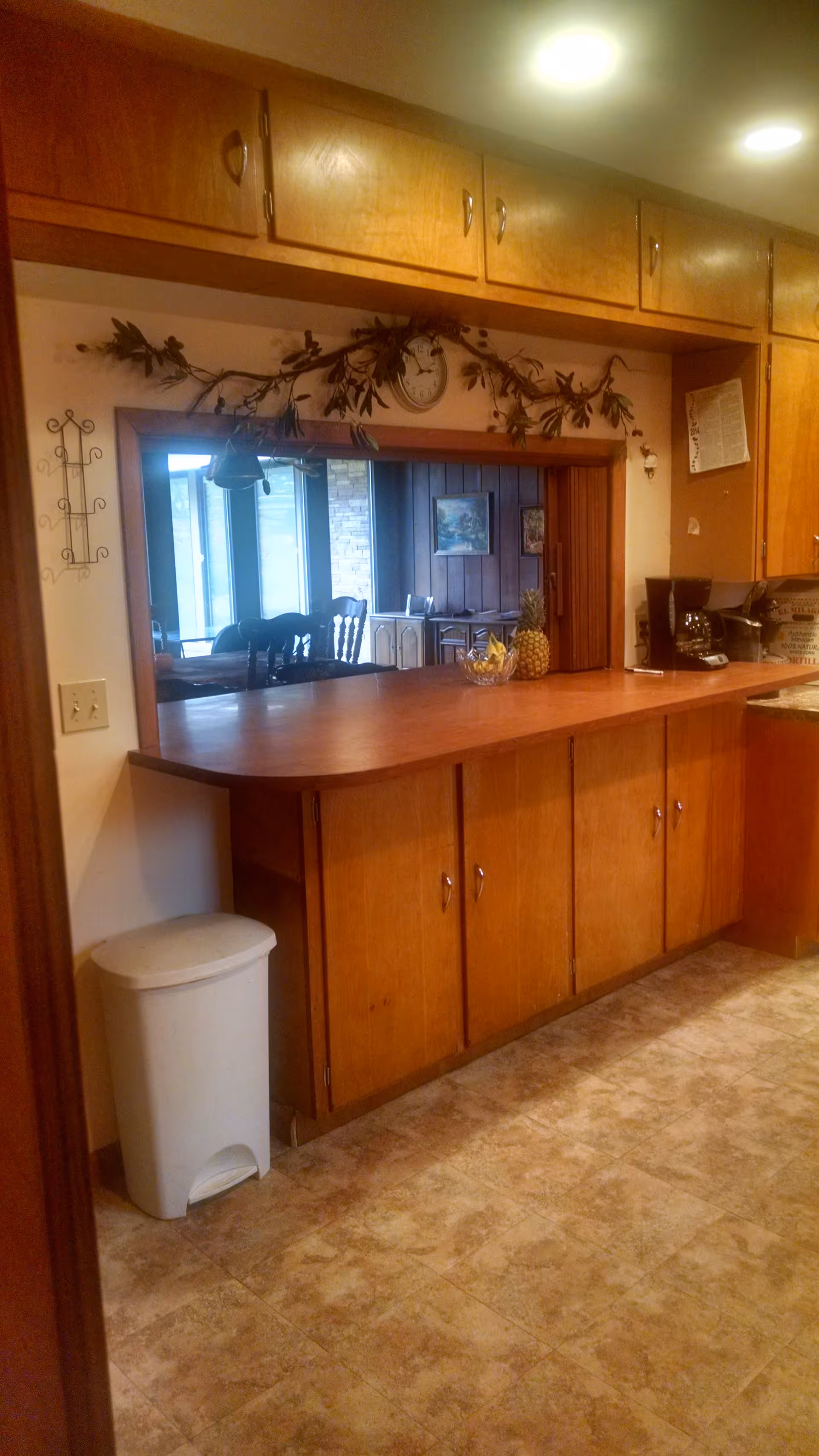 Wood-paneled kitchen pass-through with wooden cabinets, a countertop holding a fruit bowl and coffee maker, and a view into a dining area.