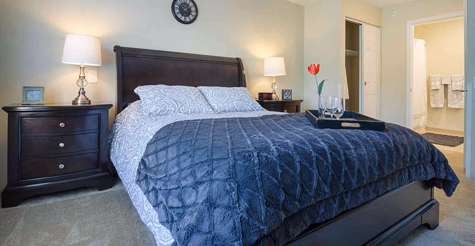 Neatly made bedroom with a dark wood bed and matching nightstands, lamps, a tray with glasses on the bed, and an open doorway to a bathroom.