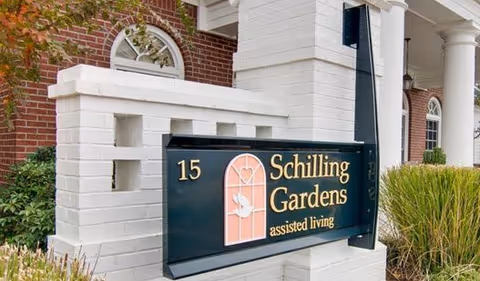 Outdoor sign for Schilling Gardens assisted living facility mounted on a white brick structure with greenery and part of a brick building in the background.