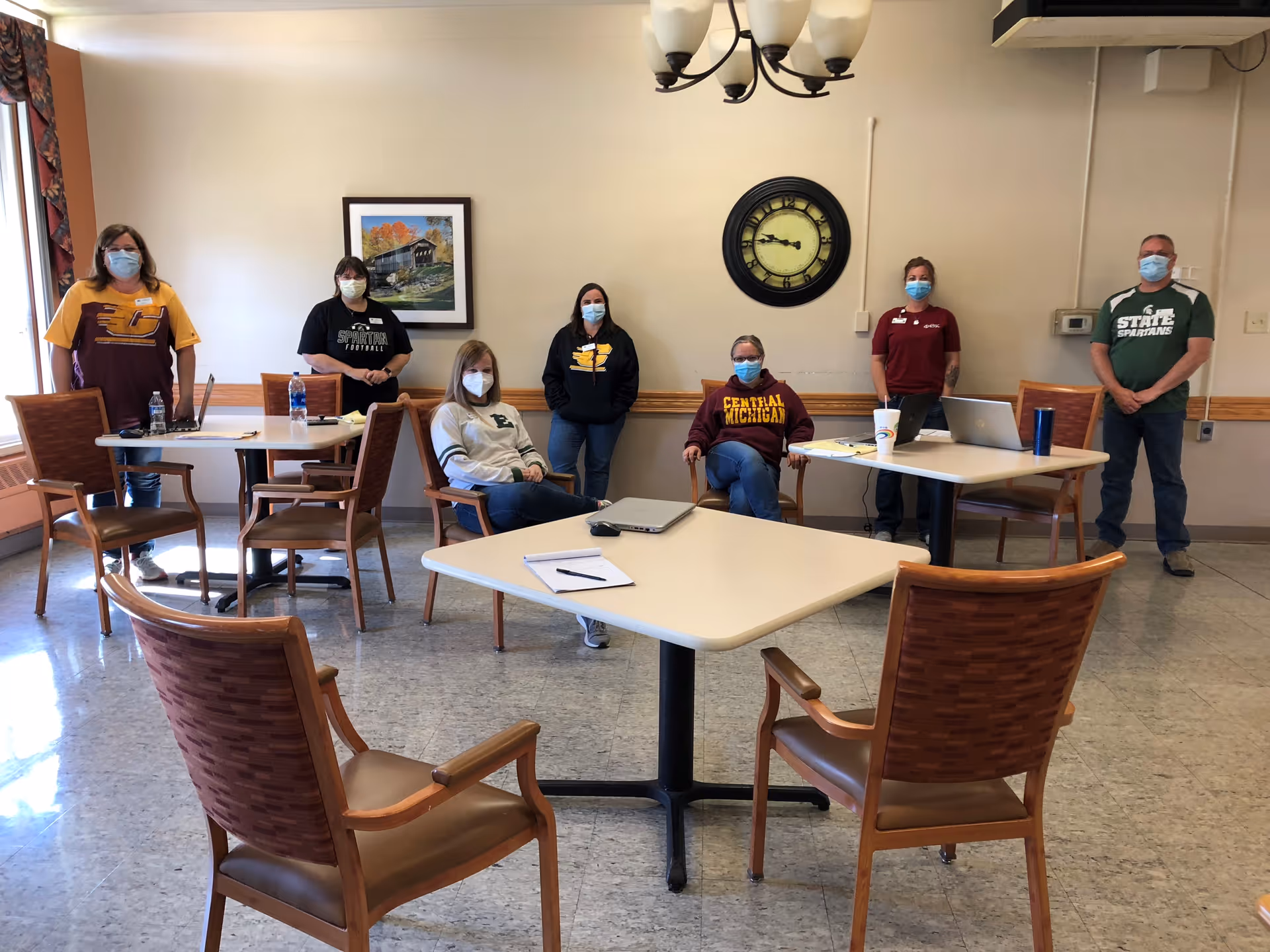 A group of seven people wearing face masks standing and sitting around tables in a room with beige walls, a large clock, and a framed picture. The people are dressed casually, some wearing college-themed sweatshirts and t-shirts. The room has several wooden chairs and a tiled floor.