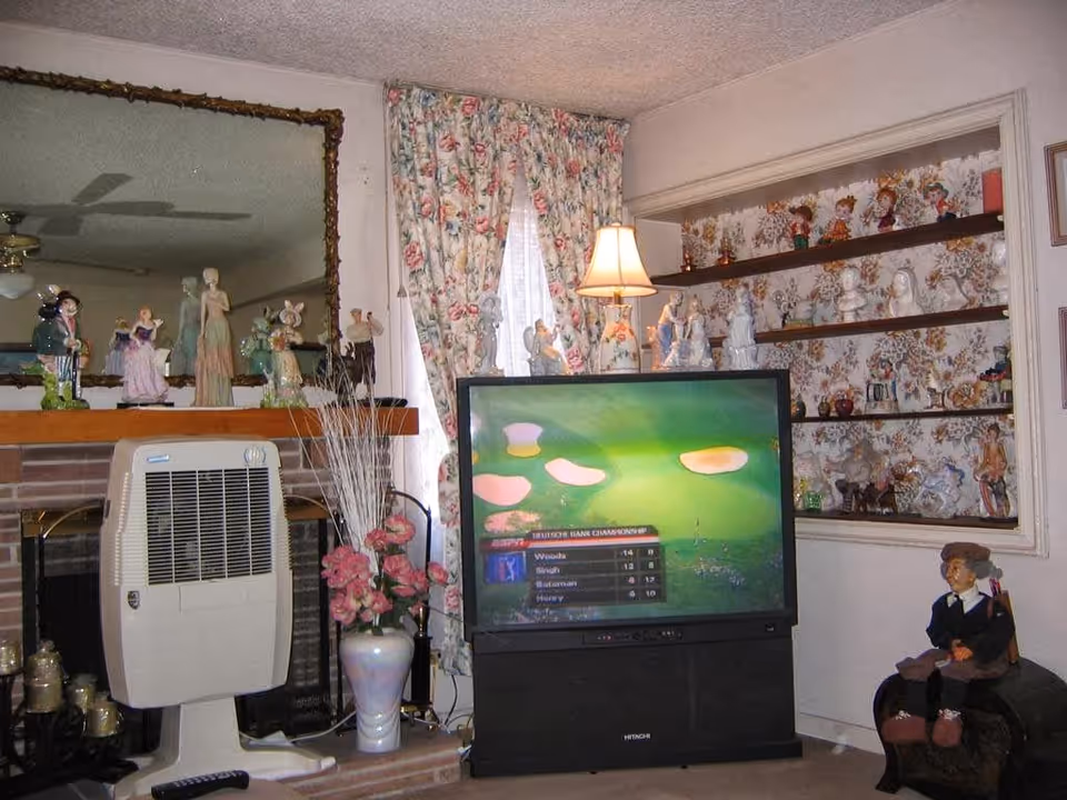 Cozy living room with a large TV, mantel and shelves filled with porcelain figurines, floral curtains and a lamp.