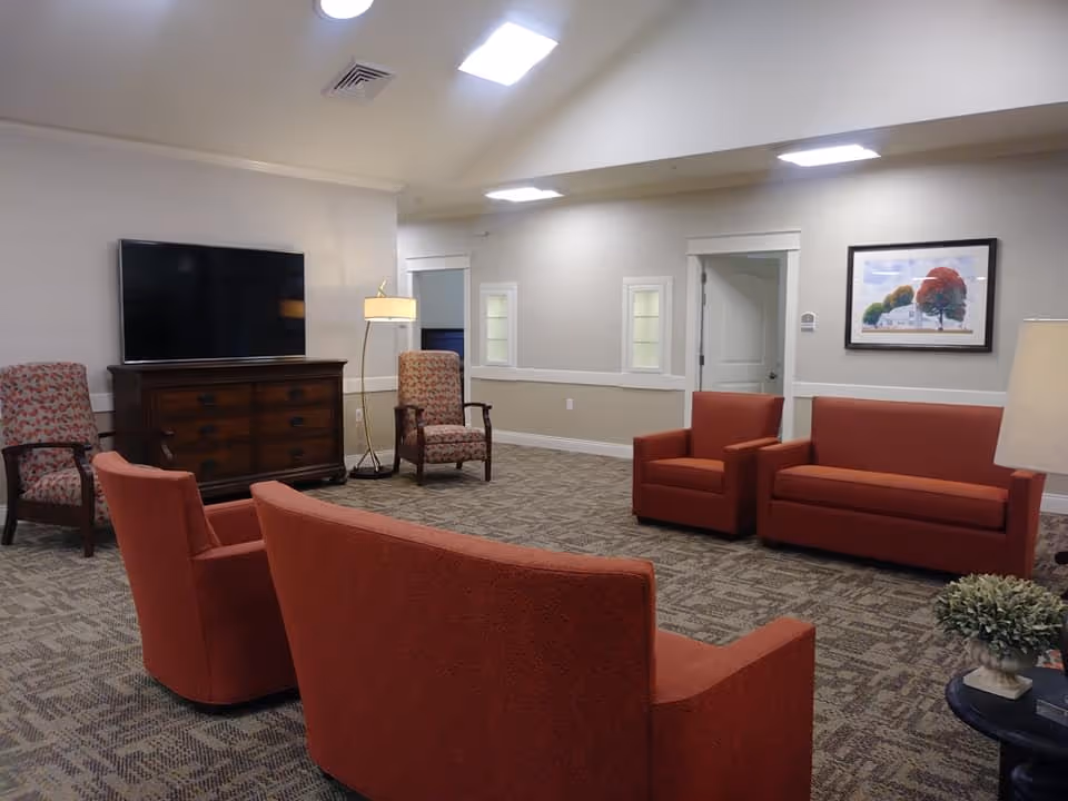 A senior living facility common area with orange upholstered chairs and sofas arranged around a carpeted floor. There is a wooden cabinet with a flat-screen TV on top, two patterned armchairs, a floor lamp, a framed picture on the wall, and a small table with a plant and books.