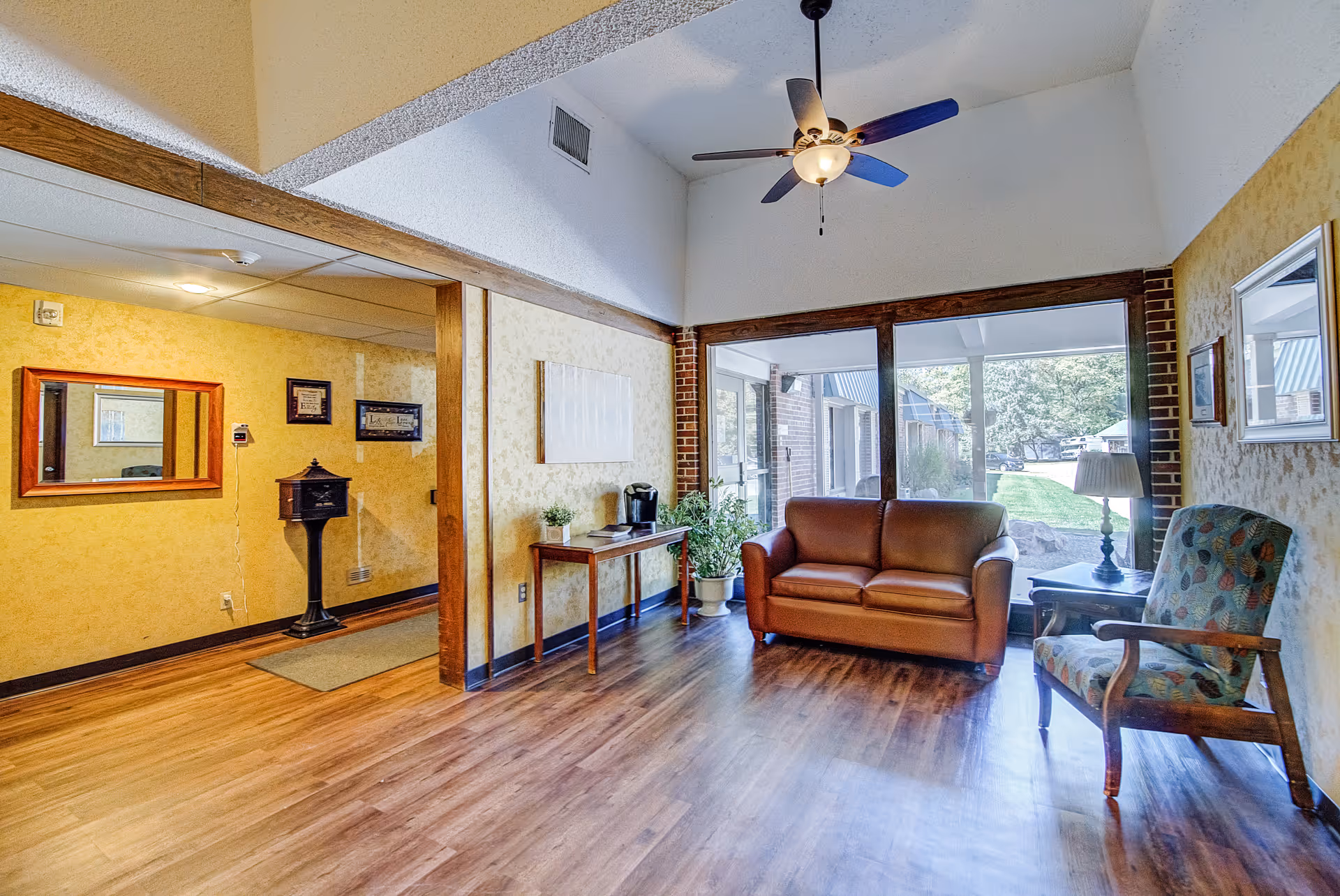 A cozy living room area in a senior living facility with a brown leather loveseat, a patterned armchair, a wooden side table with a lamp, and a small table with a coffee maker and potted plants. Large windows provide a view of the outdoor garden area. The room has wood flooring, yellow textured walls, and a ceiling fan with a light fixture.