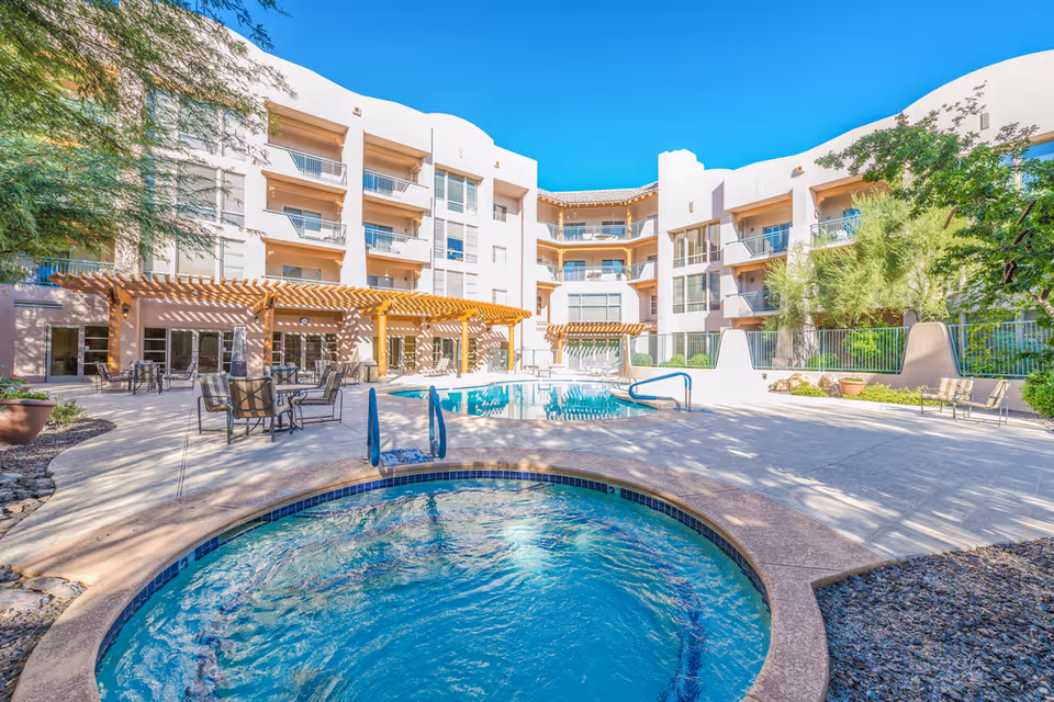 Outdoor courtyard area of a senior living facility featuring a hot tub in the foreground, a swimming pool in the middle, surrounded by patio tables and chairs, with a multi-story building in the background under a clear blue sky.